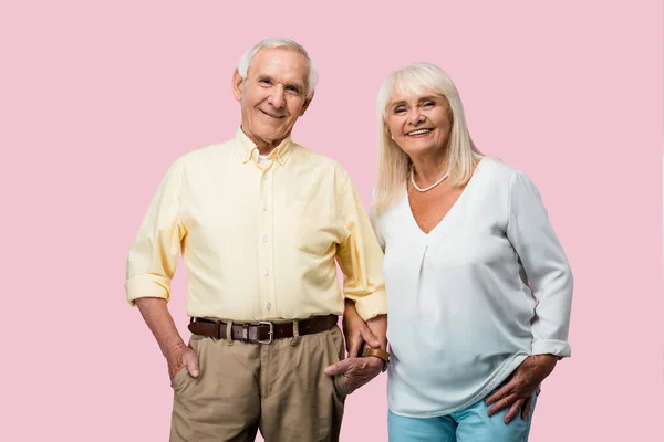 Hombre jubilado feliz con la mano en el bolsillo cogido de la mano con la esposa alegre aislado en rosa - foto de stock