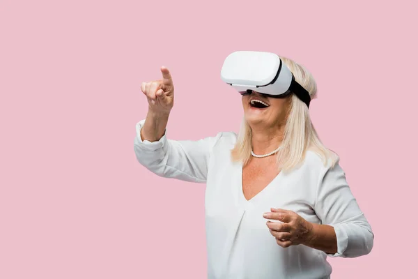 Femme retraitée pointant du doigt tout en portant un casque de réalité virtuelle isolé sur rose — Photo de stock