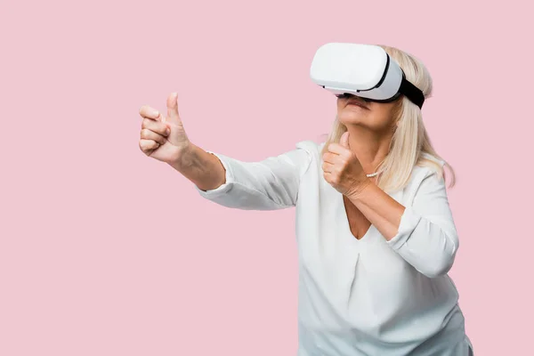 Пожилая женщина жестом в то время как носить гарнитуру виртуальной реальности изолированы на розовый — стоковое фото