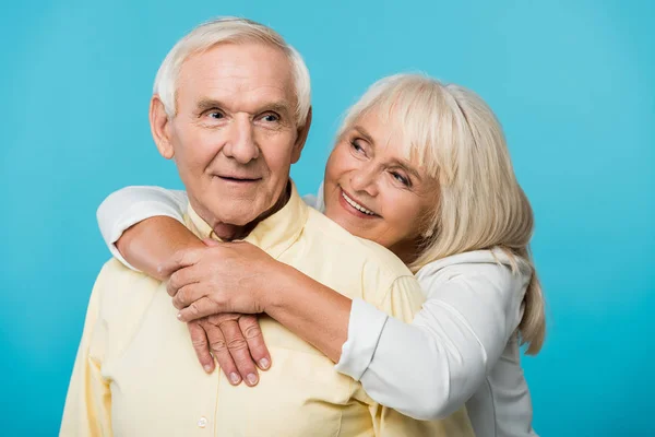 Счастливая пенсионерка обнимает веселого мужчину с седыми волосами, изолированными на голубом — стоковое фото