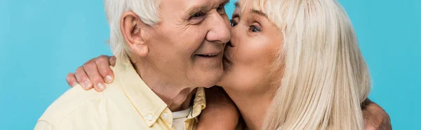 Panoramic shot of retired woman kissing cheek of cheerful man with grey hair isolated on blue — Stock Photo
