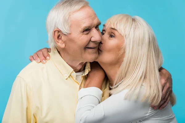 Senior woman kissing cheek of cheerful man with grey hair isolated on blue — Stock Photo