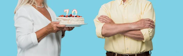 Tiro panorámico mujer jubilada sosteniendo pastel de cumpleaños cerca de marido con brazos cruzados aislados en azul - foto de stock