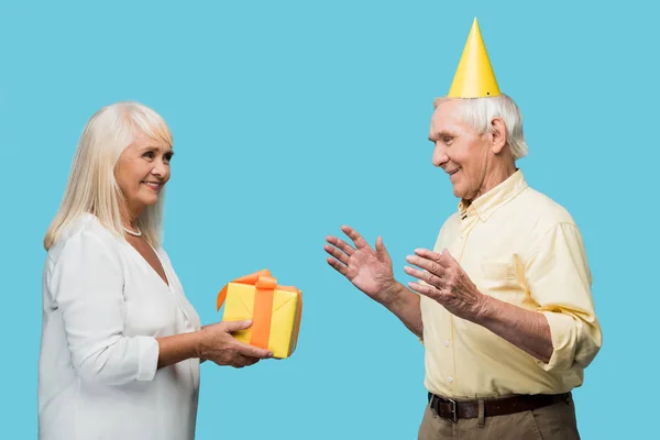 Mujer jubilada feliz dando caja de regalo amarilla a marido mayor aislado en azul - foto de stock