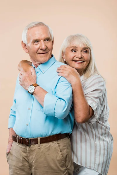 Cheerful retired woman hugging husband while holding hands isolated on beige — Stock Photo