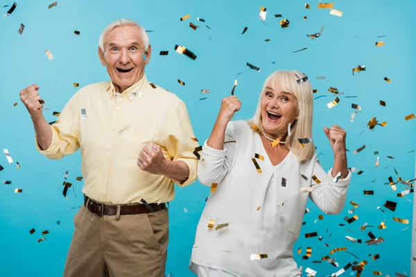 Happy retired husband and wife gesturing near falling confetti on blue — Stock Photo