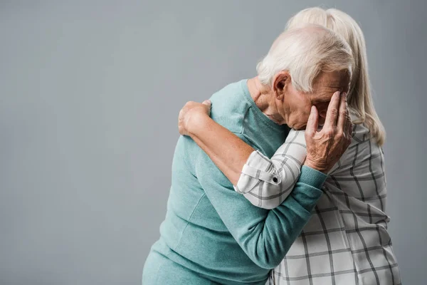 Donna anziana con i capelli grigi abbracciando sconvolto marito che copre il viso su grigio — Foto stock