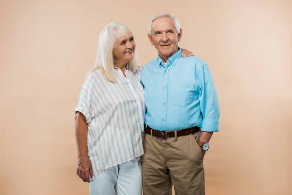 Fröhliche Rentnerin blickt Ehemann mit Hand in Tasche isoliert auf Beige stehend an — Stockfoto
