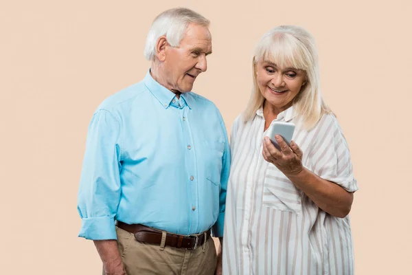 Alegre hombre retirado mirando esposa sosteniendo teléfono inteligente aislado en beige - foto de stock