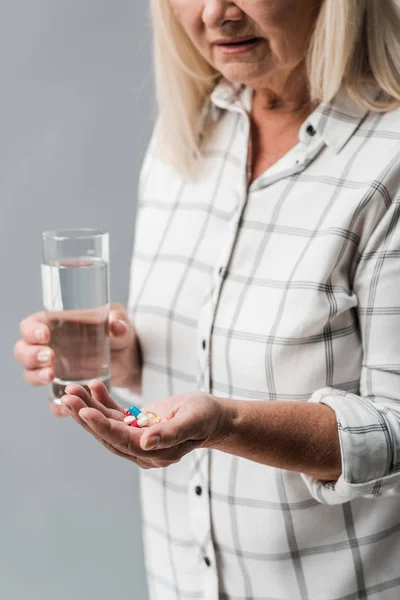 Vista recortada de la mujer mayor sosteniendo píldoras y vaso de agua aislado en gris - foto de stock