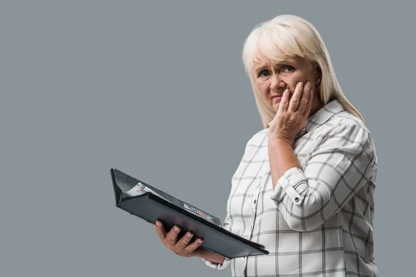 Aufgebrachte Rentnerin hält Fotoalbum in der Hand und berührt Gesicht isoliert auf Grau — Stockfoto