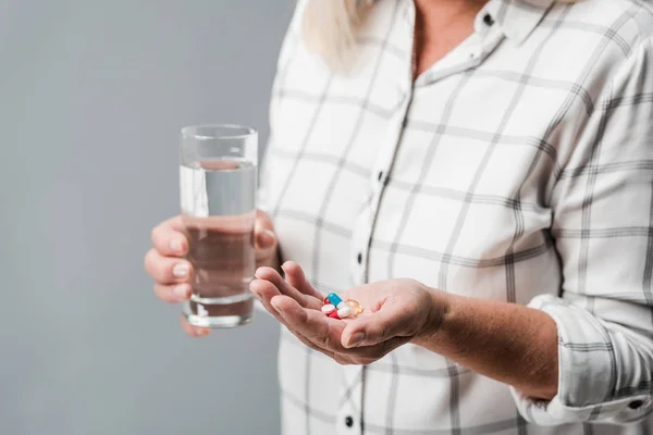 Обрезанный вид женщины на пенсии с таблетками и стаканом воды, изолированной на серой — стоковое фото