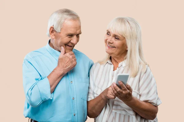 Cheerful retired woman holding smartphone and looking at husband isolated on beige — Stock Photo
