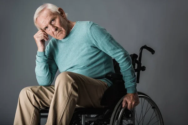 Расстроенный пенсионер, сидящий в инвалидном кресле и смотрящий в камеру на сером — стоковое фото