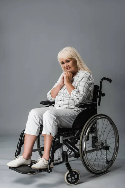 Turbato donna in pensione con le mani serrate seduto in sedia a rotelle su grigio — Foto stock