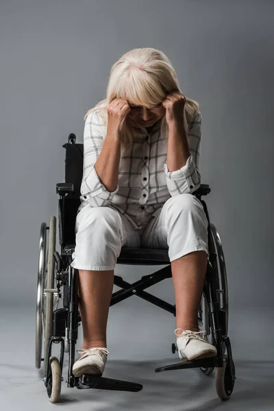 Upset retired woman sitting in wheelchair and touching head on grey — Stock Photo