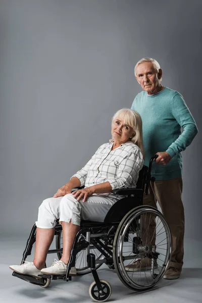 Bouleversé homme debout près de femme retraitée handicapée assis en fauteuil roulant sur gris — Photo de stock