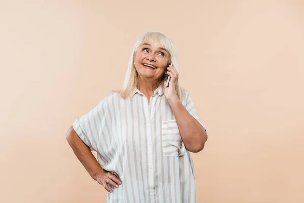 Mujer jubilada alegre de pie con la mano en la cadera y hablando en el teléfono inteligente aislado en beige - foto de stock