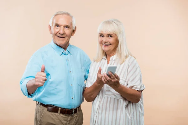 Allegra donna in pensione che tiene smartphone vicino al marito mostrando pollice in su isolato sul beige — Foto stock