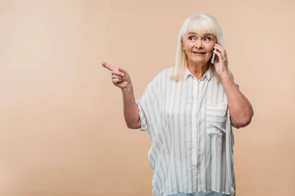 Heureuse retraitée pointant du doigt tout en parlant sur smartphone isolé sur beige — Photo de stock