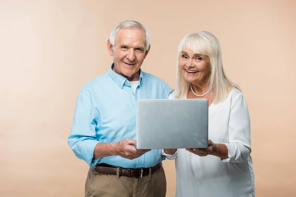 Feliz pareja jubilada usando el ordenador portátil y sonriendo aislado en beige - foto de stock