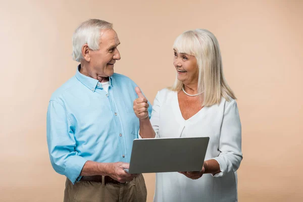 Alegre mujer jubilada mostrando el pulgar hacia arriba y mirando al marido mientras se utiliza el ordenador portátil aislado en beige - foto de stock