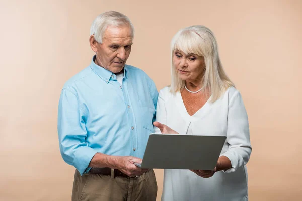 Femme retraitée gestuelle tout en regardant ordinateur portable près de mari aîné isolé sur beige — Photo de stock
