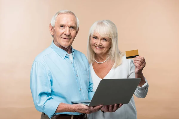 Glückliche Rentnerin mit Kreditkarte in der Nähe ihres älteren Mannes und Laptop isoliert auf beige — Stockfoto