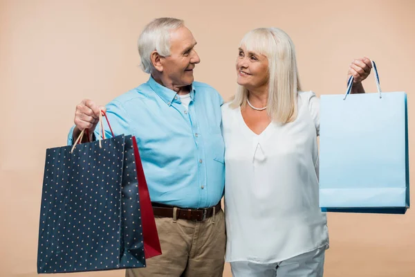 Cheerful retired couple looking at each other and holding shopping bags isolated on beige — Stock Photo