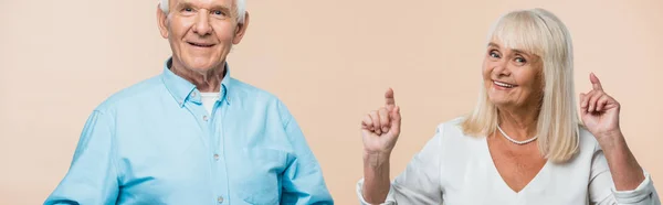Tiro panorámico de mujer jubilada feliz señalando con los dedos cerca del marido aislado en beige - foto de stock
