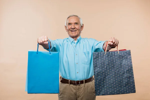 Cheerful retired man smiling while holding shopping bags on beige — Stock Photo