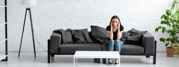 Mujer joven deprimida sentada en un sofá en una habitación espaciosa y cogida de la mano cerca de la cabeza - foto de stock