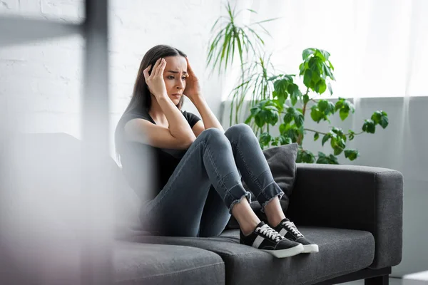 Selective focus of young woman suffering from depression while sitting on couch and holding hands near head — Stock Photo