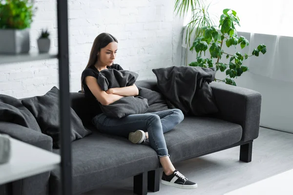 Mujer joven deprimida sentada en el sofá en casa y abrazando la almohada - foto de stock