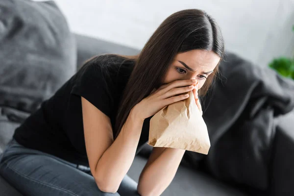 Young woman breathing into paper bag while suffering from panic attack at home — Stock Photo