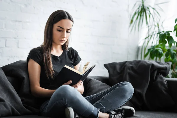 Grave jovem mulher ler livro enquanto sentado no sofá com pernas cruzadas — Fotografia de Stock