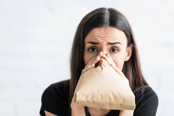 Scared woman breathing into paper bag and looking at camera whule suffering from panick attact at home — Stock Photo