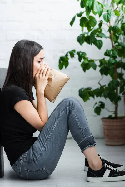 Visão lateral da jovem mulher sentada no chão e respirando em saco de papel enquanto sofre de ataque de pânico — Fotografia de Stock