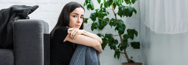 Панорамний знімок розчарованої молодої жінки, яка страждає від депресії, сидячи на підлозі вдома і дивлячись на камеру — стокове фото
