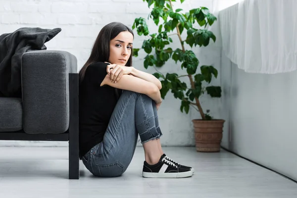 Mujer joven deprimida sentada en el suelo en casa y mirando a la cámara - foto de stock