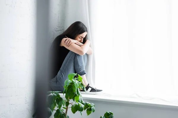 Depressed young woman crying while sitting on window sill and hiding face in crossed arms — Stock Photo