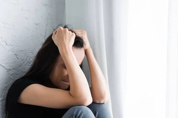 Mujer joven que sufre de depresión y llanto mientras está sentada junto a la ventana y cogida de la mano cerca de la cabeza - foto de stock