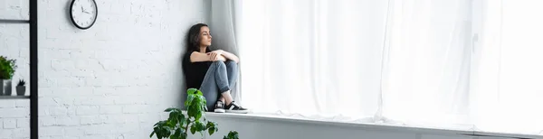Plano panorámico de mujer joven deprimida sentada en el alféizar de la ventana con los brazos cruzados y mirando hacia otro lado - foto de stock