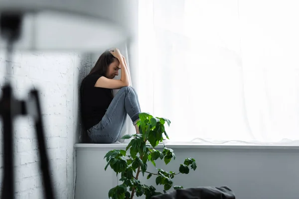 Enfoque selectivo de la mujer joven deprimida llorando mientras está sentado en el alféizar de la ventana y cogido de la mano en la cabeza - foto de stock