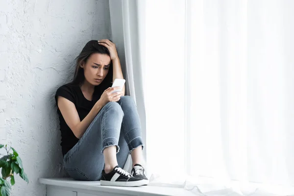 Jovem deprimida sentada na soleira da janela em casa e usando smartphone — Fotografia de Stock