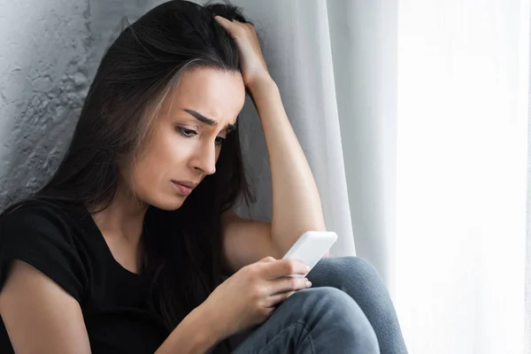 Chica decepcionada usando el teléfono inteligente mientras se sienta junto a la ventana en casa y sosteniendo la mano en la cabeza - foto de stock