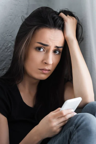 Depressive junge Frau nutzt Smartphone, hält Hand auf Kopf und blickt in Kamera — Stockfoto