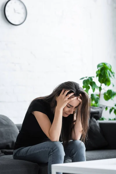 Депрессивная девушка плачет, сидя дома на диване и держа руки за голову — стоковое фото