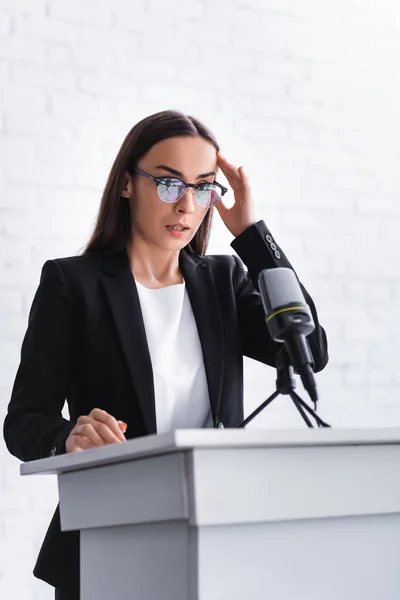 Young lecturer suffering from fear of public speaking standing on podium tribune and holding hand near head — Stock Photo