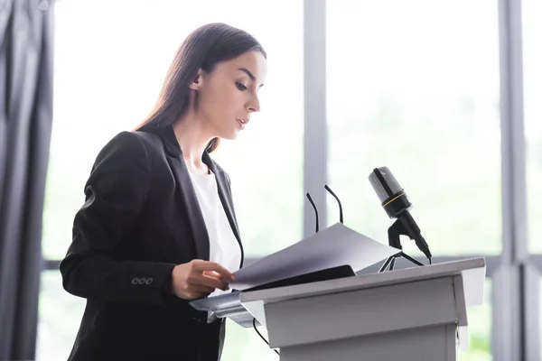 Attractive, attentive lecturer standing on podium tribune and looking at documents — Stock Photo